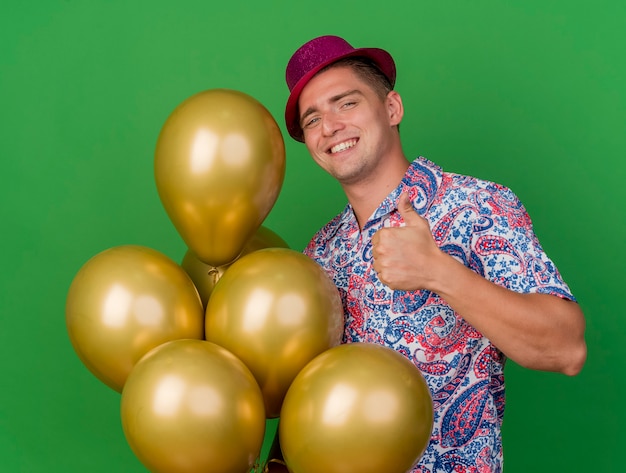 Sonriente joven fiestero con sombrero rosa sosteniendo globos mostrando el pulgar hacia arriba aislado en verde