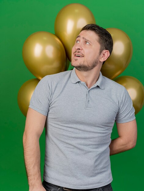 Sonriente joven fiestero vistiendo camisa gris de pie delante de globos aislados en verde