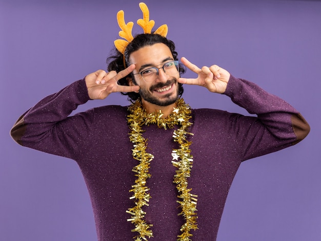Sonriente joven guapo con aro de pelo de navidad con guirnalda en el cuello mostrando gesto de paz aislado sobre fondo azul.