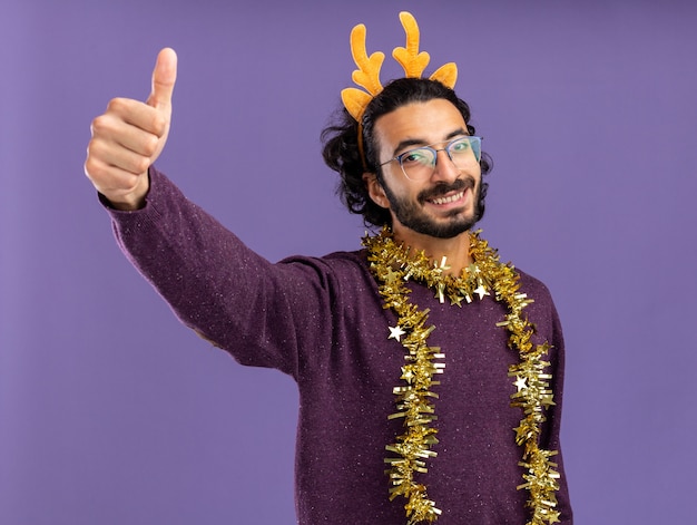 Sonriente joven guapo con aro de pelo de Navidad con guirnalda en el cuello mostrando el pulgar hacia arriba aislado sobre fondo azul.