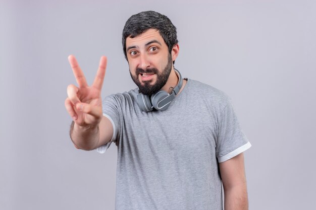 Sonriente joven guapo con auriculares en el cuello y haciendo el signo de la paz aislado en la pared blanca