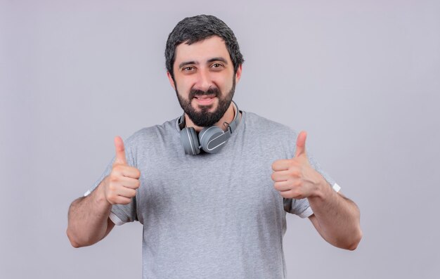 Sonriente joven guapo con auriculares en el cuello y mostrando los pulgares para arriba aislado en la pared blanca