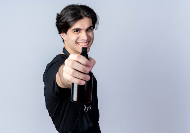 Sonriente joven guapo barbero en uniforme sosteniendo una botella de spray en la cámara