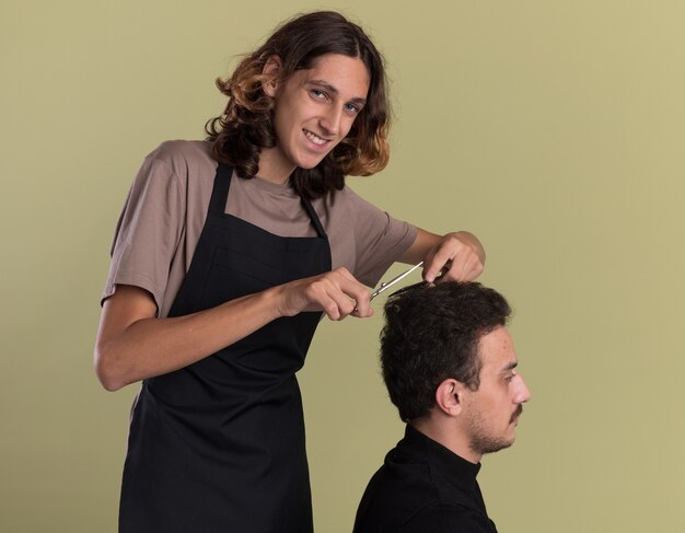 Sonriente joven guapo barbero vistiendo uniforme haciendo corte de pelo para su joven cliente aislado en la pared verde oliva