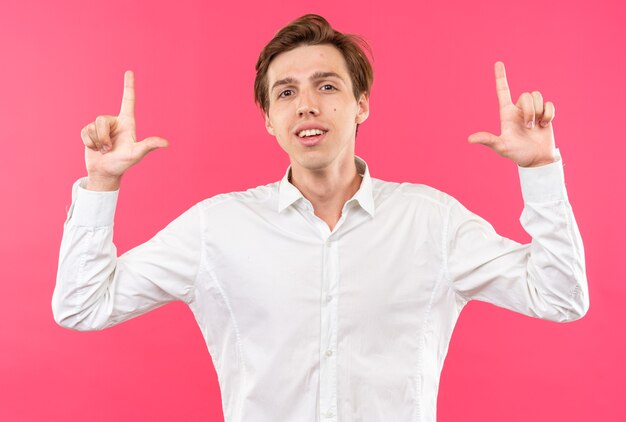 Sonriente joven guapo con camisa blanca apunta hacia arriba aislado en la pared rosa