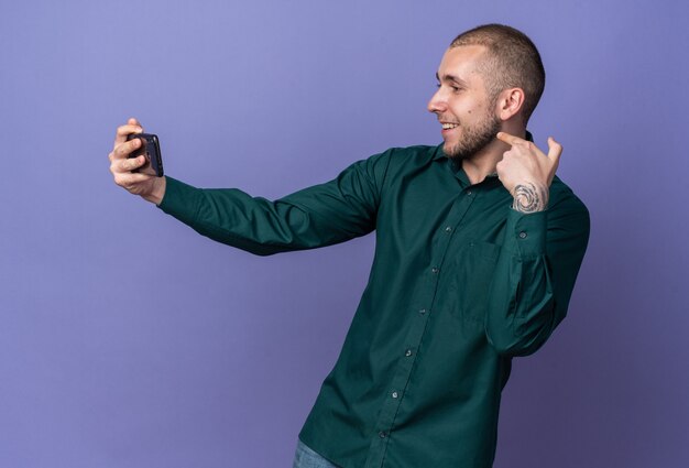 Sonriente joven guapo con camisa verde tomar un selfie apunta a sí mismo