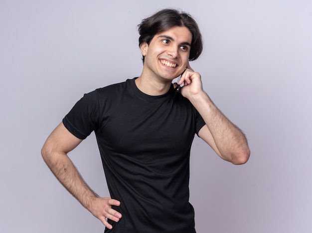 Sonriente joven guapo con camiseta negra habla por teléfono poniendo la mano en la cadera aislada en la pared blanca