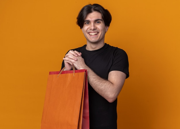 Sonriente joven guapo con camiseta negra sosteniendo el bolso aislado en la pared naranja