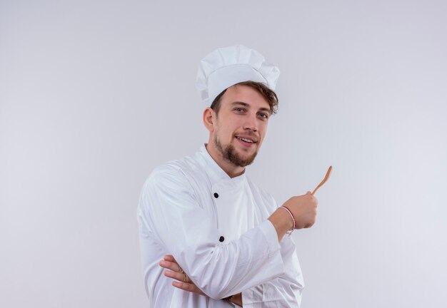 Un sonriente joven y guapo chef barbudo hombre vestido con uniforme de cocina blanco y sombrero sosteniendo una cuchara de madera mientras mira en una pared blanca