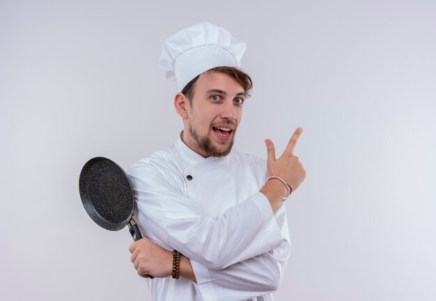 Un sonriente joven guapo chef barbudo con uniforme blanco de cocina y sombrero apuntando hacia arriba y sosteniendo una sartén mientras mira en una pared blanca