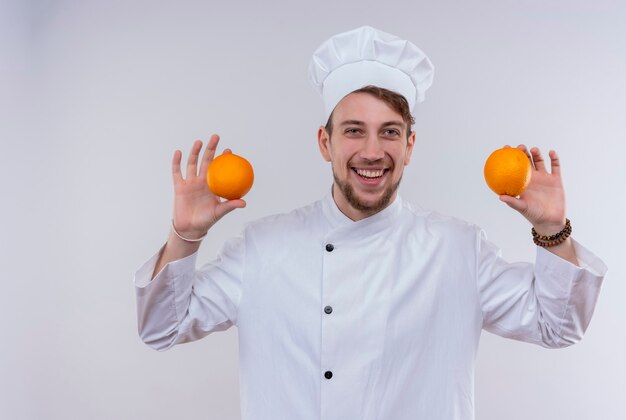 Un sonriente joven guapo chef barbudo vestido con uniforme de cocina blanco y sombrero sosteniendo naranjas mientras mira en una pared blanca