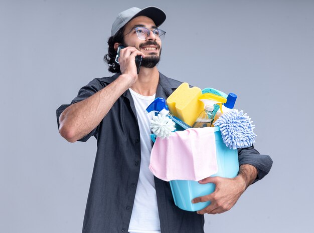 Sonriente joven guapo chico de limpieza con camiseta y gorra sosteniendo un cubo de herramientas de limpieza y habla por teléfono aislado en la pared blanca