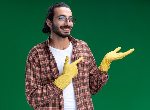 Sonriente joven guapo chico de limpieza con camiseta y guantes fingiendo sostener y apunta a algo aislado en la pared verde con espacio de copia