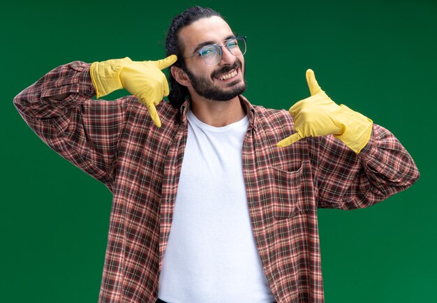 Sonriente joven guapo chico de limpieza con camiseta y guantes que muestran gesto de llamada telefónica aislado en la pared verde