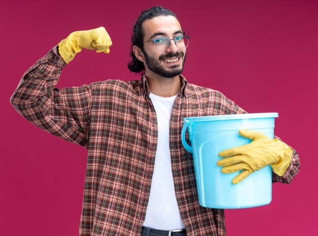 Sonriente joven guapo chico de limpieza con camiseta y guantes sosteniendo un cubo que muestra un gesto fuerte aislado en la pared rosa