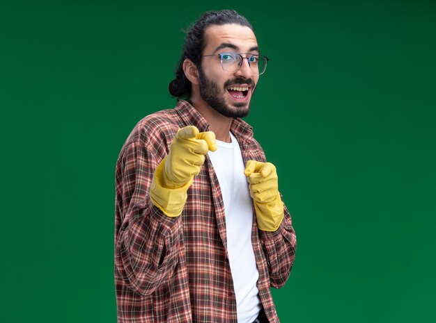 Sonriente joven guapo chico de limpieza con camiseta y puntos de guantes en el frente aislado en la pared verde con espacio de copia