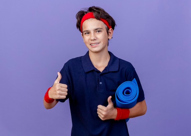 Sonriente joven guapo deportivo con diadema y muñequeras con aparatos dentales sosteniendo estera de yoga mostrando los pulgares para arriba aislados en la pared púrpura con espacio de copia