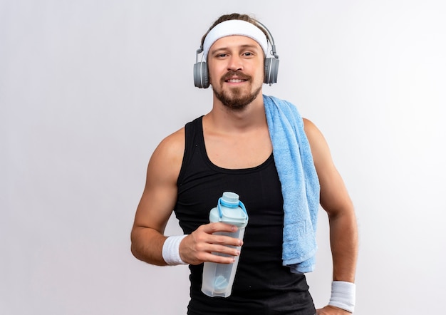 Sonriente joven guapo deportivo con diadema y muñequeras y auriculares sosteniendo una botella de agua con una toalla en el hombro aislado en el espacio en blanco