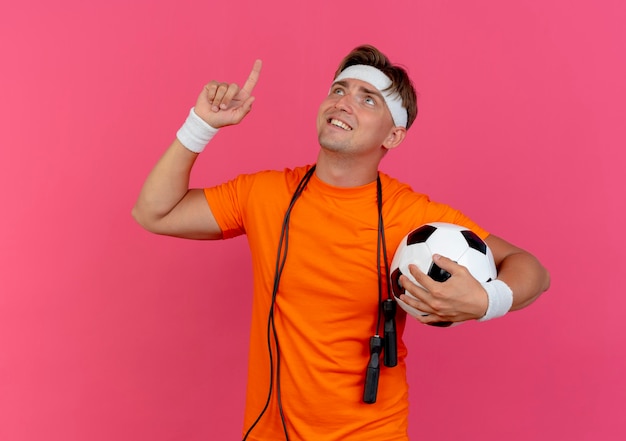 Sonriente joven guapo deportivo con diadema y muñequeras con saltar la cuerda alrededor del cuello sosteniendo un balón de fútbol mirando y apuntando hacia arriba aislado en la pared rosa