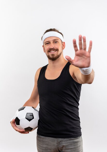 Sonriente joven guapo deportivo con diadema y muñequeras sosteniendo un balón de fútbol y estirando la mano aislada en el espacio en blanco