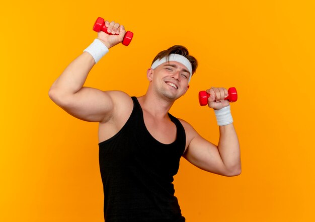 Sonriente joven guapo deportivo con diadema y muñequeras sosteniendo pesas aisladas en la pared naranja