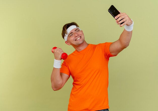 Sonriente joven guapo deportivo con diadema y muñequeras sosteniendo teléfono móvil y mancuernas tomando selfie aislado en pared verde oliva