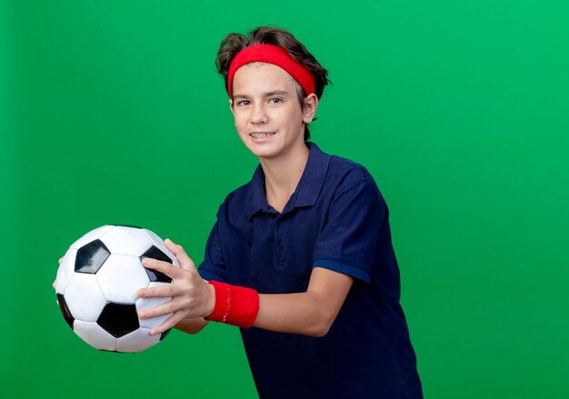 Sonriente joven guapo deportivo vistiendo diadema y muñequeras con aparatos dentales estirando el balón de fútbol aislado en la pared verde con espacio de copia