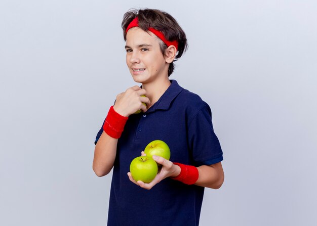 Sonriente joven guapo deportivo vistiendo diadema y muñequeras con aparatos dentales mirando recto sosteniendo manzanas aisladas sobre fondo blanco con espacio de copia