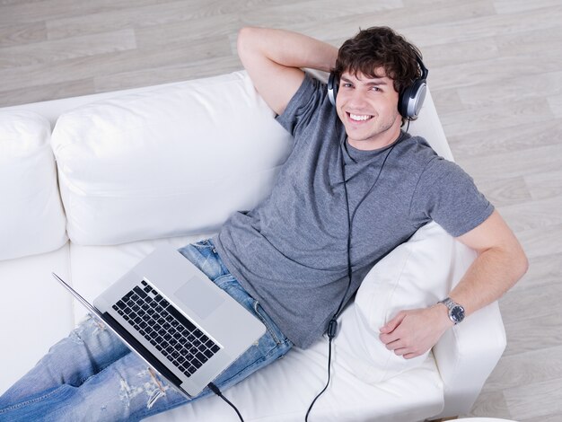 Sonriente joven guapo escuchando música en auriculares desde el portátil - ángulo alto