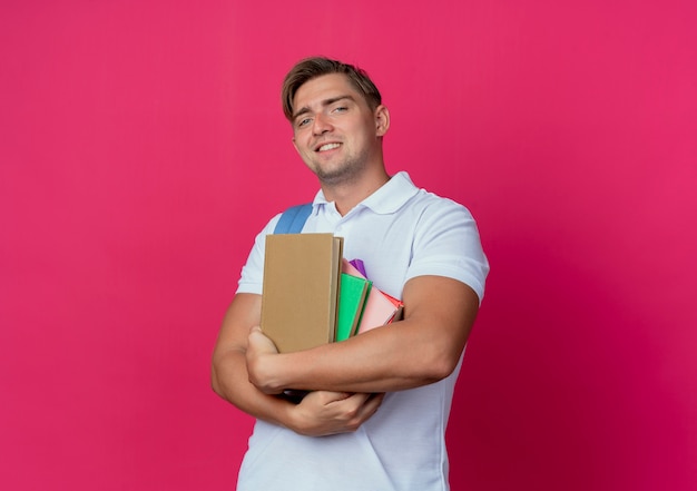 Sonriente joven guapo estudiante sosteniendo libros aislados en rosa