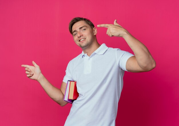 Sonriente joven guapo estudiante sosteniendo libros y puntos al lado aislado en rosa