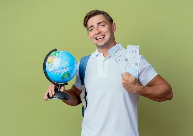 Sonriente joven guapo estudiante vistiendo bolsa trasera sosteniendo boletos y globo aislado en verde oliva