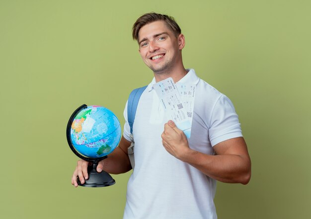 Sonriente joven guapo estudiante vistiendo bolsa trasera sosteniendo boletos y globo aislado en verde oliva