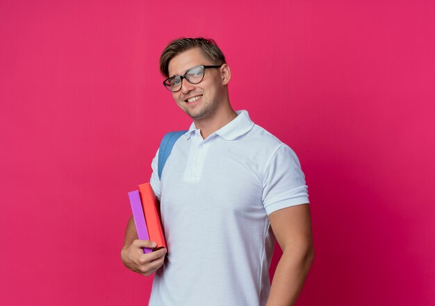 Sonriente joven guapo estudiante vistiendo bolsa trasera sosteniendo libros aislados en rosa