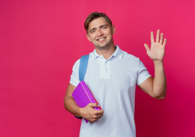 Sonriente joven guapo estudiante vistiendo bolsa trasera sosteniendo libros y mostrando gesto de saludo aislado en la pared rosa