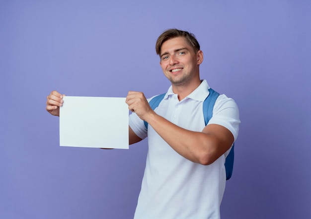 Sonriente joven guapo estudiante vistiendo bolsa trasera sosteniendo papel aislado en azul