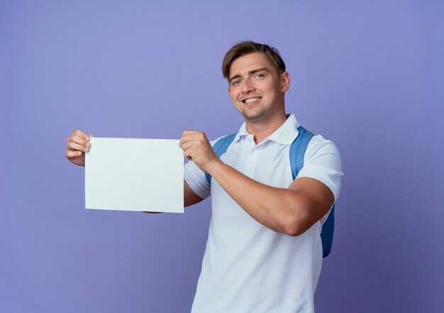 Sonriente joven guapo estudiante vistiendo bolsa trasera sosteniendo papel aislado en azul