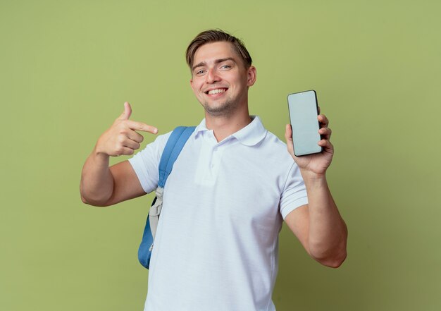 Sonriente joven guapo estudiante vistiendo bolsa trasera sosteniendo y puntos en el teléfono aislado en verde oliva