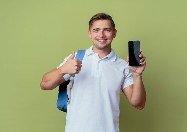 Sonriente joven guapo estudiante vistiendo bolsa trasera sosteniendo el teléfono con el pulgar hacia arriba aislado sobre fondo verde oliva