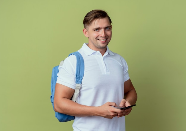 Sonriente joven guapo estudiante vistiendo bolso trasero sosteniendo teléfono aislado en verde oliva