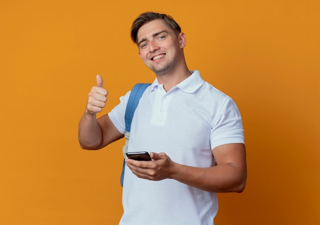 Sonriente joven guapo estudiante vistiendo bolso trasero sosteniendo teléfono y su pulgar arriba aislado en naranja