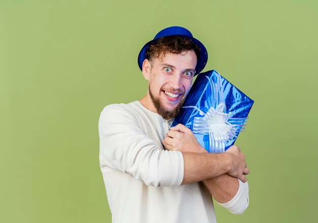 Sonriente joven guapo partido eslavo con sombrero de fiesta con caja de regalo mirando a cámara aislada sobre fondo verde oliva con espacio de copia