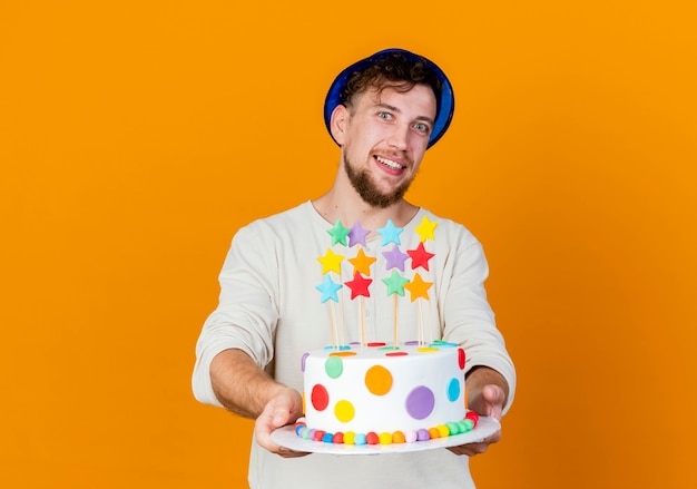 Sonriente joven guapo partido eslavo con sombrero de fiesta estirando la torta de cumpleaños con estrellas y mirando a cámara aislada sobre fondo naranja con espacio de copia