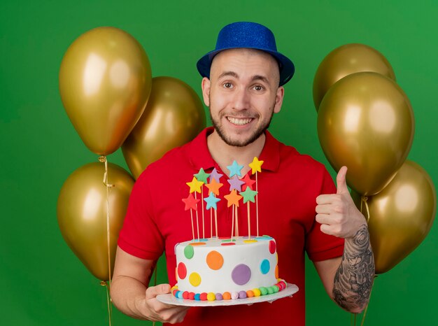 Sonriente joven guapo partido eslavo con sombrero de fiesta de pie delante de globos sosteniendo pastel de cumpleaños mostrando el pulgar hacia arriba mirando a cámara aislada sobre fondo verde