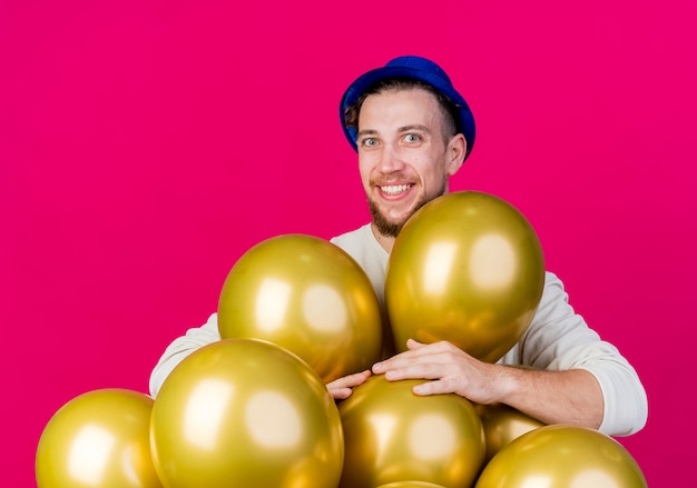 Sonriente joven guapo partido eslavo con sombrero de fiesta de pie detrás de globos mirando a cámara aislada sobre fondo carmesí