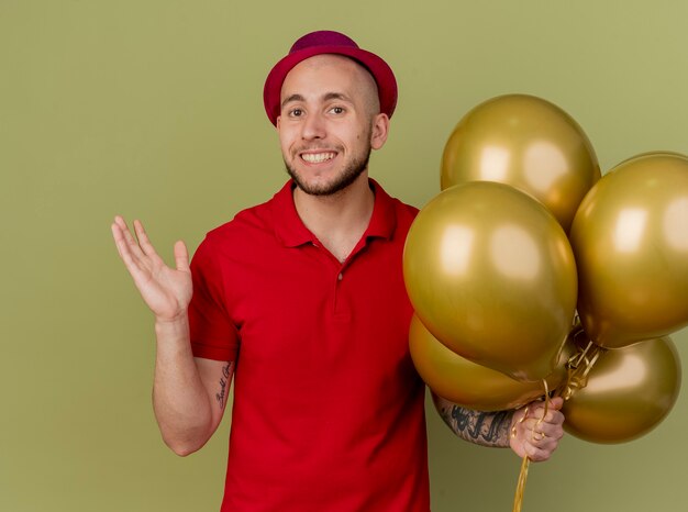 Sonriente joven guapo partido eslavo con sombrero de fiesta sosteniendo globos mirando a la cámara mostrando la mano vacía aislada sobre fondo verde oliva