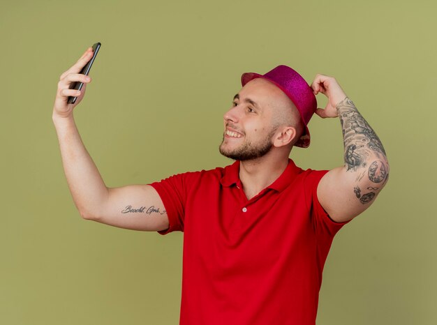 Sonriente joven guapo partido eslavo con sombrero de fiesta tocando el sombrero y tomando selfie aislado sobre fondo verde oliva