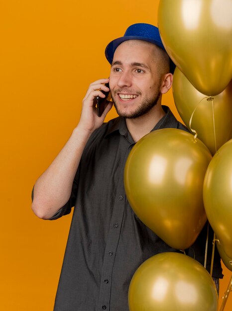 Sonriente joven guapo partido eslavo vistiendo gorro de fiesta sosteniendo globos hablando por teléfono mirando directamente aislado sobre fondo naranja