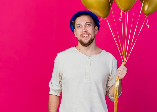 Sonriente joven guapo partido eslavo vistiendo gorro de fiesta sosteniendo globos mirando al frente aislado en la pared rosa con espacio de copia