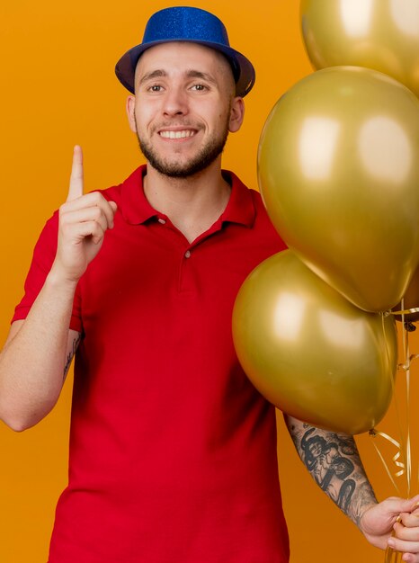 Sonriente joven guapo partido eslavo vistiendo gorro de fiesta sosteniendo globos mirando a la cámara apuntando hacia arriba aislado sobre fondo naranja